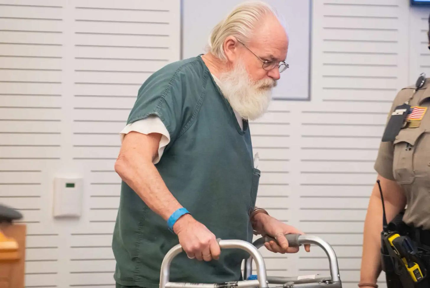 Michael Royce Sparks, white hair and a long beard, holding a walker and wearing detention center scrubs, walks in the courtroom past the bailiff.