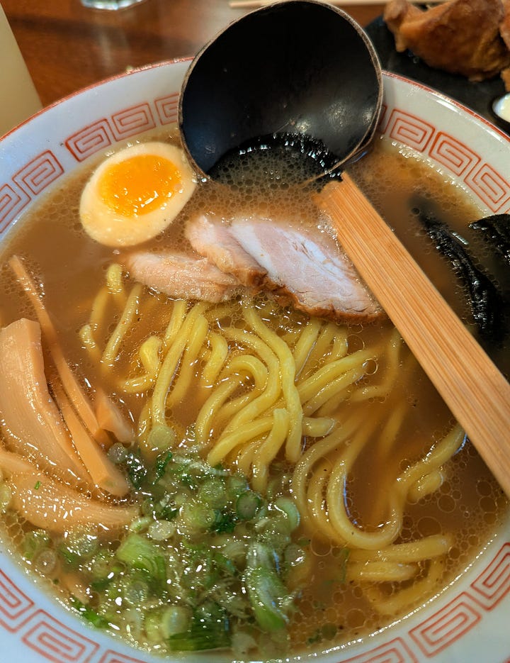 Two ramens. On the left featured noodles, bamboo, green onions, egg, and pork with a long spoon. On the right, a deeper brown/red broth with minced meat. 