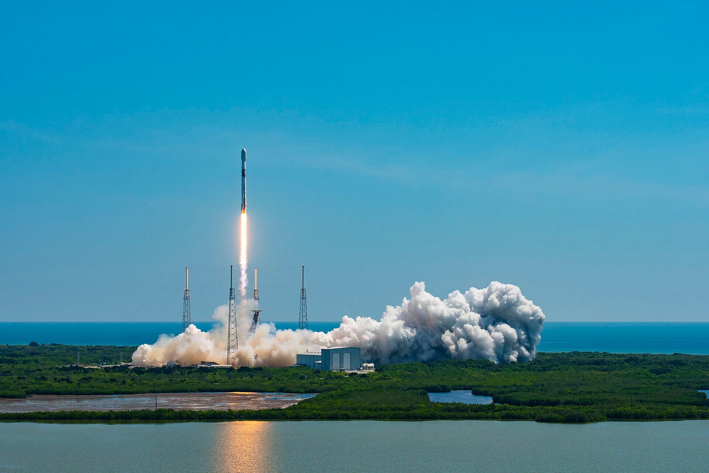 Against a clear blue sky, the Falcon 9 Rocket has just lifted off the ground. A jet of flame is visible in the rocket's wake, and great plumes of smoke billow out across the surrounding land.