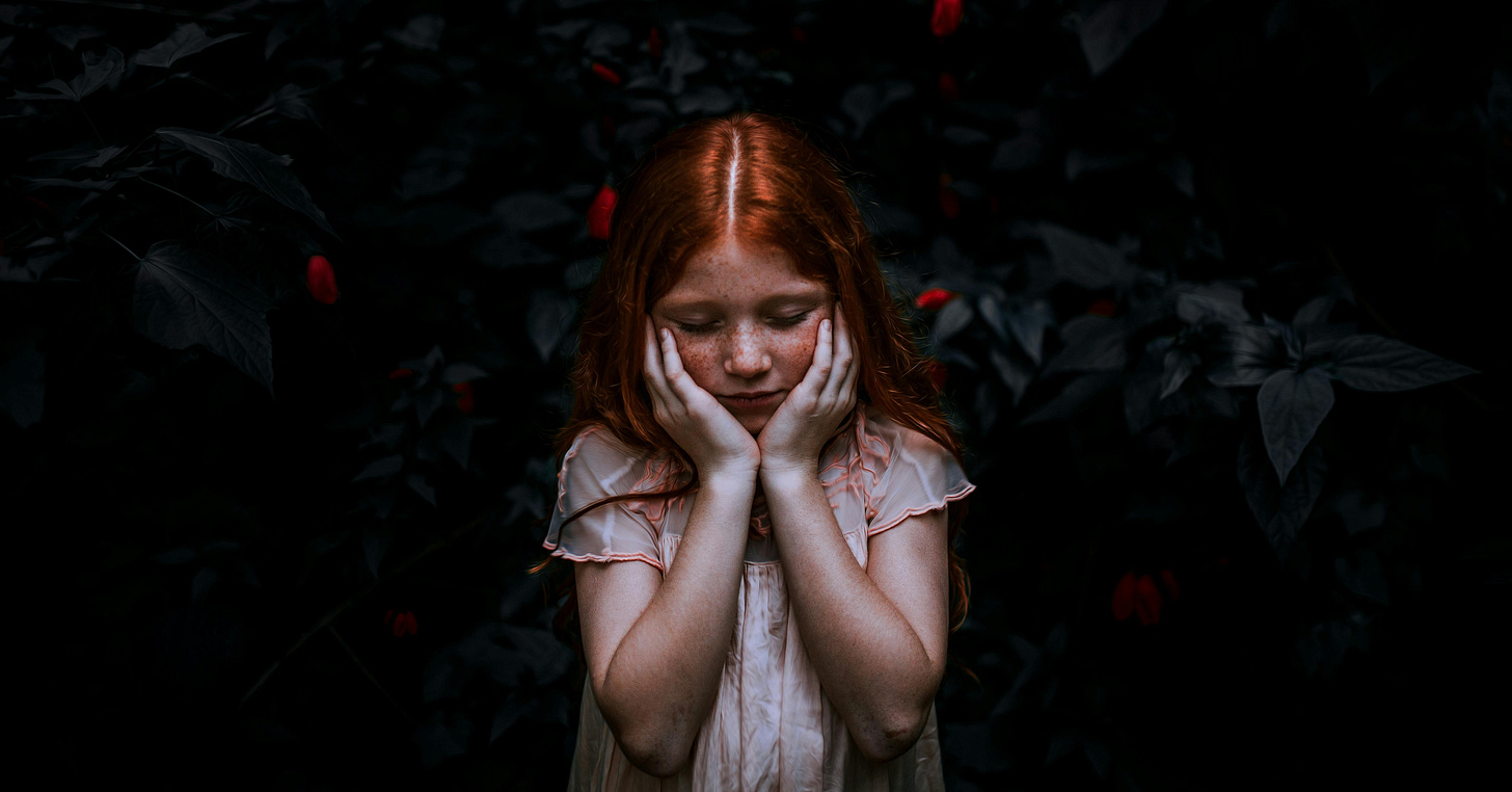 A young girl with long red hair stands against a dark backdrop, holding her hands close to her chest, surrounded by red flowers.