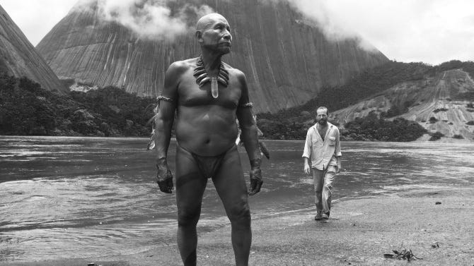 Black and white film still showing an indigenous elder wearing ritual ornaments walking beside a river in the rainforest, followed by a man in modern clothing; a scene from Embrace of the Serpent, symbolizing the meeting of ancient and modern wisdom