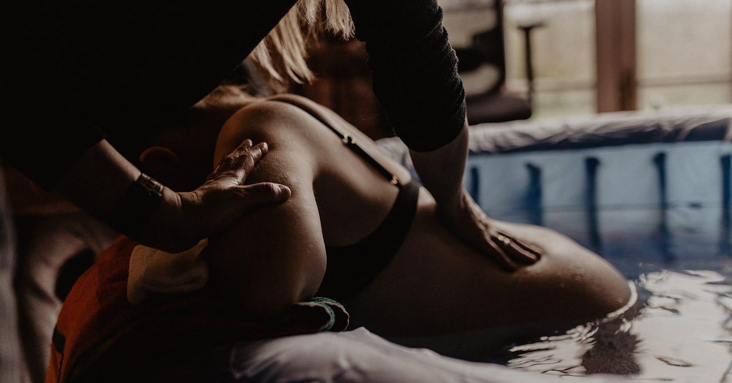 A close-up of a woman in a birthing pool, receiving support and massage during labor from a caregiver, creating a serene atmosphere.