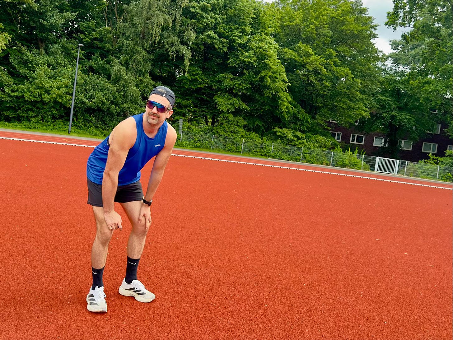 A person bending forward with hands on their knees on a new red running track, catching their breath after a run, trees visible in the background.