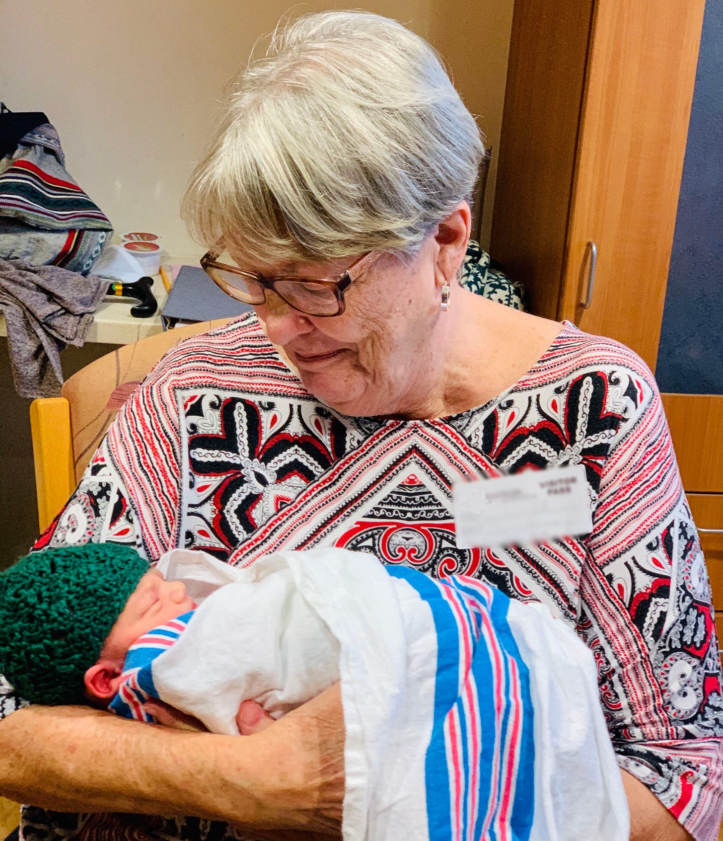 A woman with white hair and glasses, wearing a red, white, and black patterned shirt, holds a newborn baby.