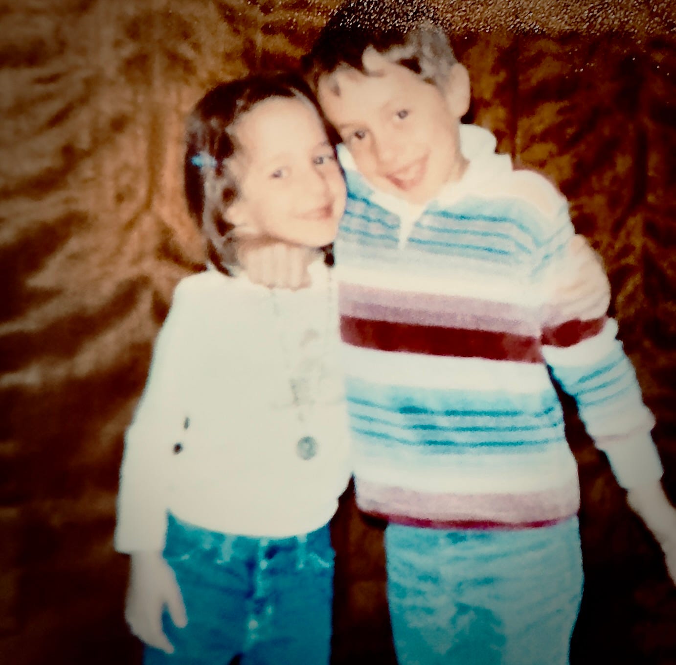 Vintage color photo of Angela and her brother Tony as young children, arms around each other, cheek to cheek, both smiling. Tony wears a blue and white striped sweater; Angela wears a white jacket and jeans. A textured brown pantry door is behind them. Vintage color photo of Angela and her brother Tony as young children, arms around each other, cheek to cheek, both smiling. Tony wears a blue and white striped sweater; Angela wears a white jacket and jeans. A textured brown pantry door is behind them.