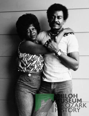 A man and woman stand close together, embracing in front of a textured wall. The woman wears a patterned top and jeans, the man a light shirt and jeans. Both look directly at the camera, conveying a sense of warmth and intimacy. Text reads: 