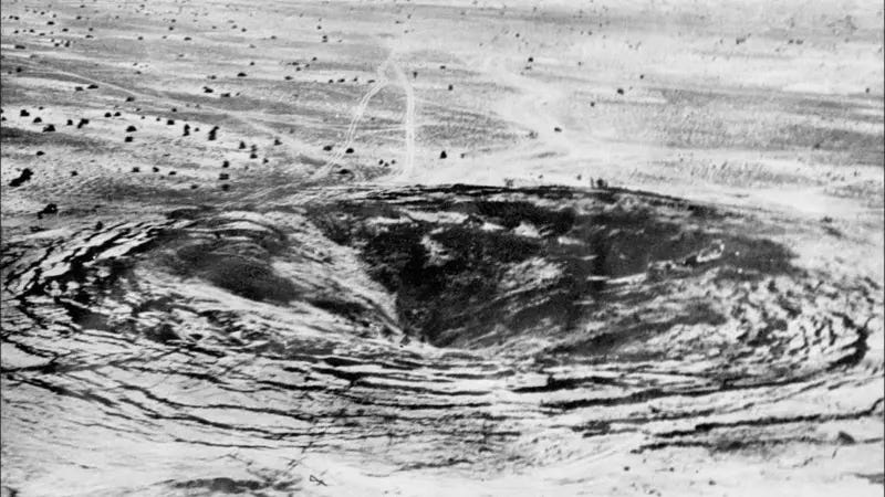 Aerial black and white view of a broad, shallow crater with radial ridges in flat desert scrubland, with vehicle tracks running past it