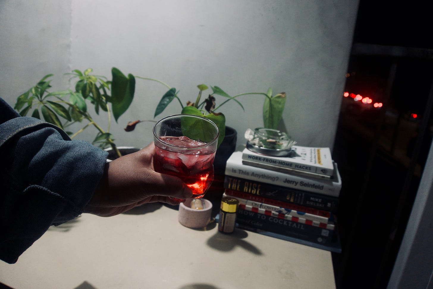 Hand holding a glass of red drink over a dimly lit tabletop with potted plants and a small stack of books, city lights glowing softly in the background. Hand holding a glass of red drink over a dimly lit tabletop with potted plants and a small stack of books, city lights glowing softly in the background.