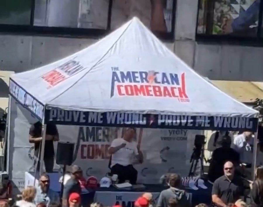 A tent with a white canopy displaying "The American Comeback" and "PROVE ME WRONG" text in red and blue. Charlie Kirk stands under the tent, wearing a white shirt and holding a baseball bat. People are gathered around, some wearing red hats, with a stage and speakers visible.