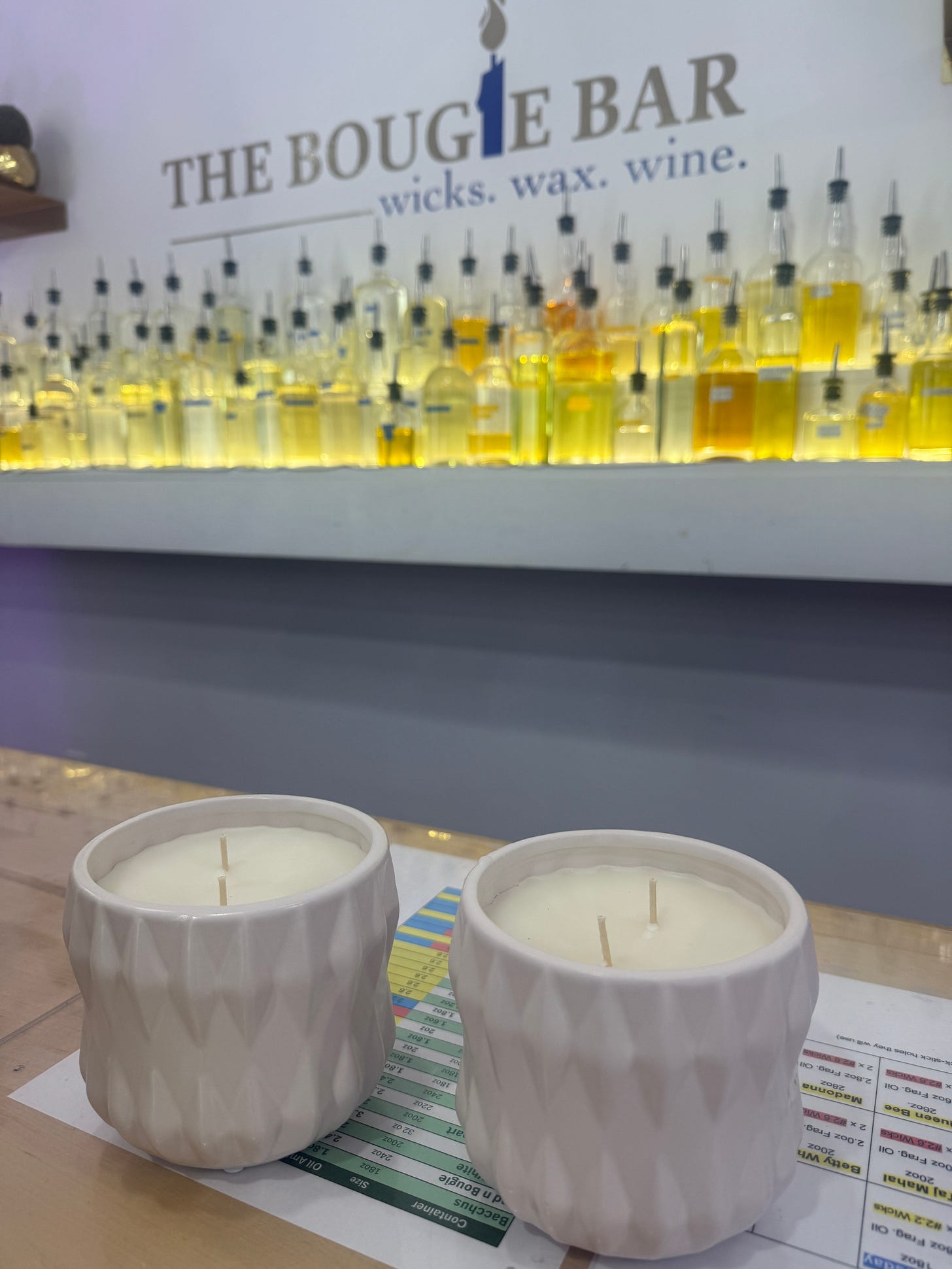 Two white ceramic candles with textured geometric pattern and white wicks, displayed on a counter with yellow fragrance oils visible in the background at The Bougie Bar Two white ceramic candles with textured geometric pattern and white wicks, displayed on a counter with yellow fragrance oils visible in the background at The Bougie Bar