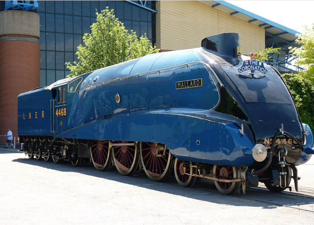 A blue steam locomotive named Mallard, with its number 4468 visible, parked outside a building. The train features distinctive streamlined design and large wheels.
