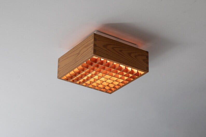 May include: A rectangular wooden ceiling light fixture with a grid pattern on the bottom. The fixture is made of light-colored wood with visible grain and is mounted on a white ceiling. The grid emits a warm, orange glow. May include: A rectangular wooden ceiling light fixture with a grid pattern on the bottom. The fixture is made of light-colored wood with visible grain and is mounted on a white ceiling. The grid emits a warm, orange glow.