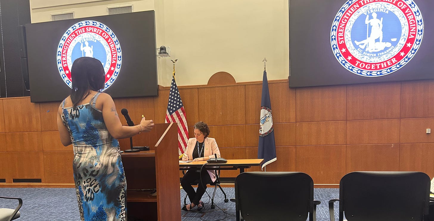 A woman wearing a dress has her back to the camera as she speaks to another woman seated behind a table.