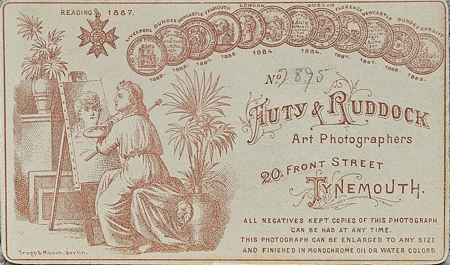 Vintage card from Fluty & Ruddock, art photographers, featuring an illustration of a female artist painting a portrait on an easel. The card includes decorative elements, text detailing the photographers' address, and various awards.