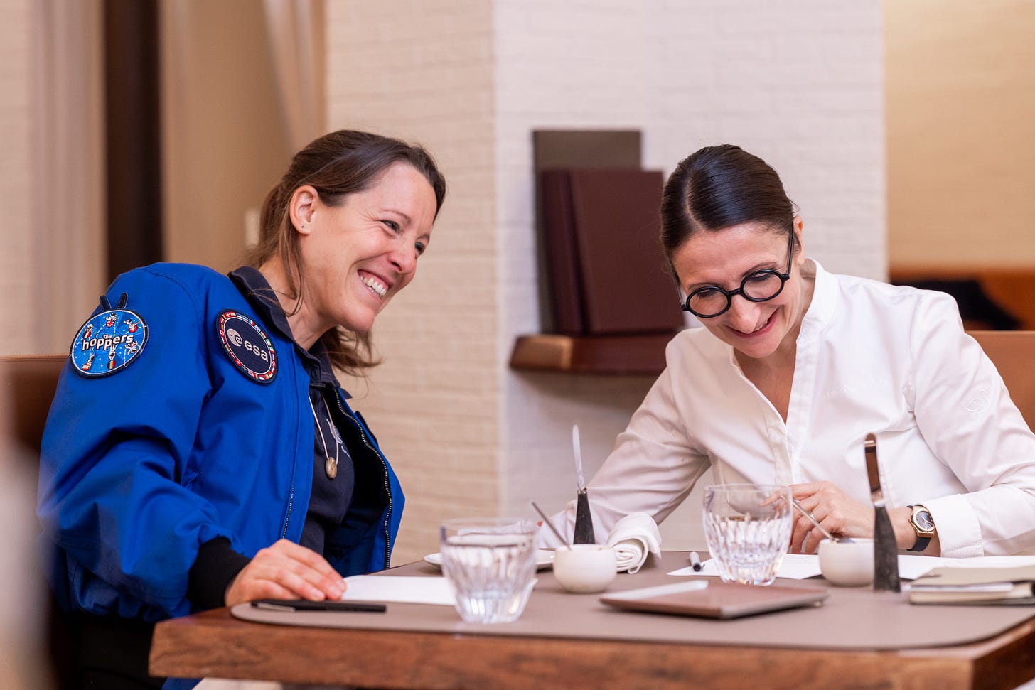ESA astronaut Sophie Adenot, with her blue ESA jacket adorned with mission patches, is laughing with Anne-Sophie Pic as the pair sit at a restaurant table with water glasses, notebooks and pens. ESA astronaut Sophie Adenot, with her blue ESA jacket adorned with mission patches, is laughing with Anne-Sophie Pic as the pair sit at a restaurant table with water glasses, notebooks and pens.
