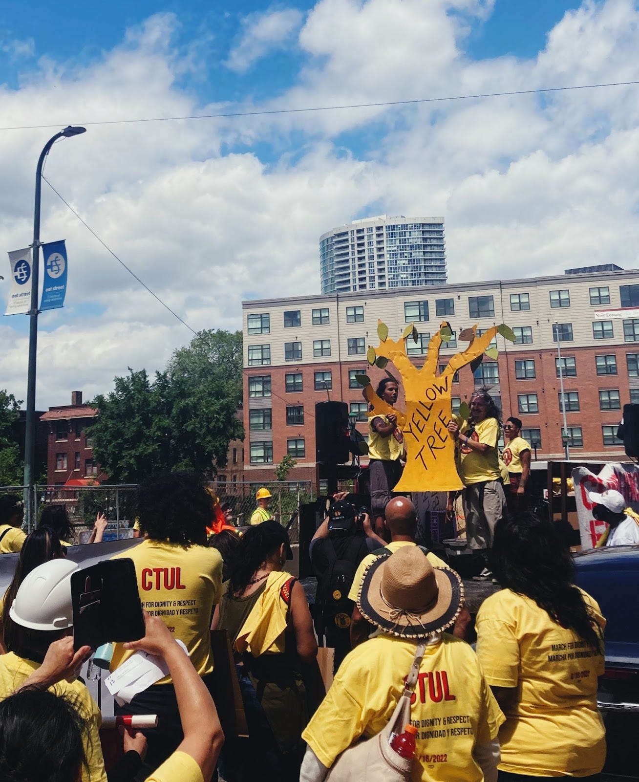 a crowd of people in yellow shirts stands behind a group of people standing on a truck holding a yellow cardboard tree with the words "yellow tree" on it a crowd of people in yellow shirts stands behind a group of people standing on a truck holding a yellow cardboard tree with the words "yellow tree" on it