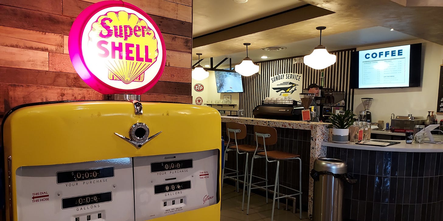 A close-up of a vintage Super Shell gasoline pump is lit up in warm light. To the right, in the background is a barista pulling a shot at an espresso machine behind a tile-faced counter and bar. A close-up of a vintage Super Shell gasoline pump is lit up in warm light. To the right, in the background is a barista pulling a shot at an espresso machine behind a tile-faced counter and bar.