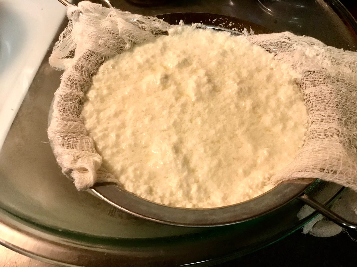 A strainer lined with cheesecloth and filled with the beginnings of ricotta — curdled milk with the liquids draining into the bowl below
