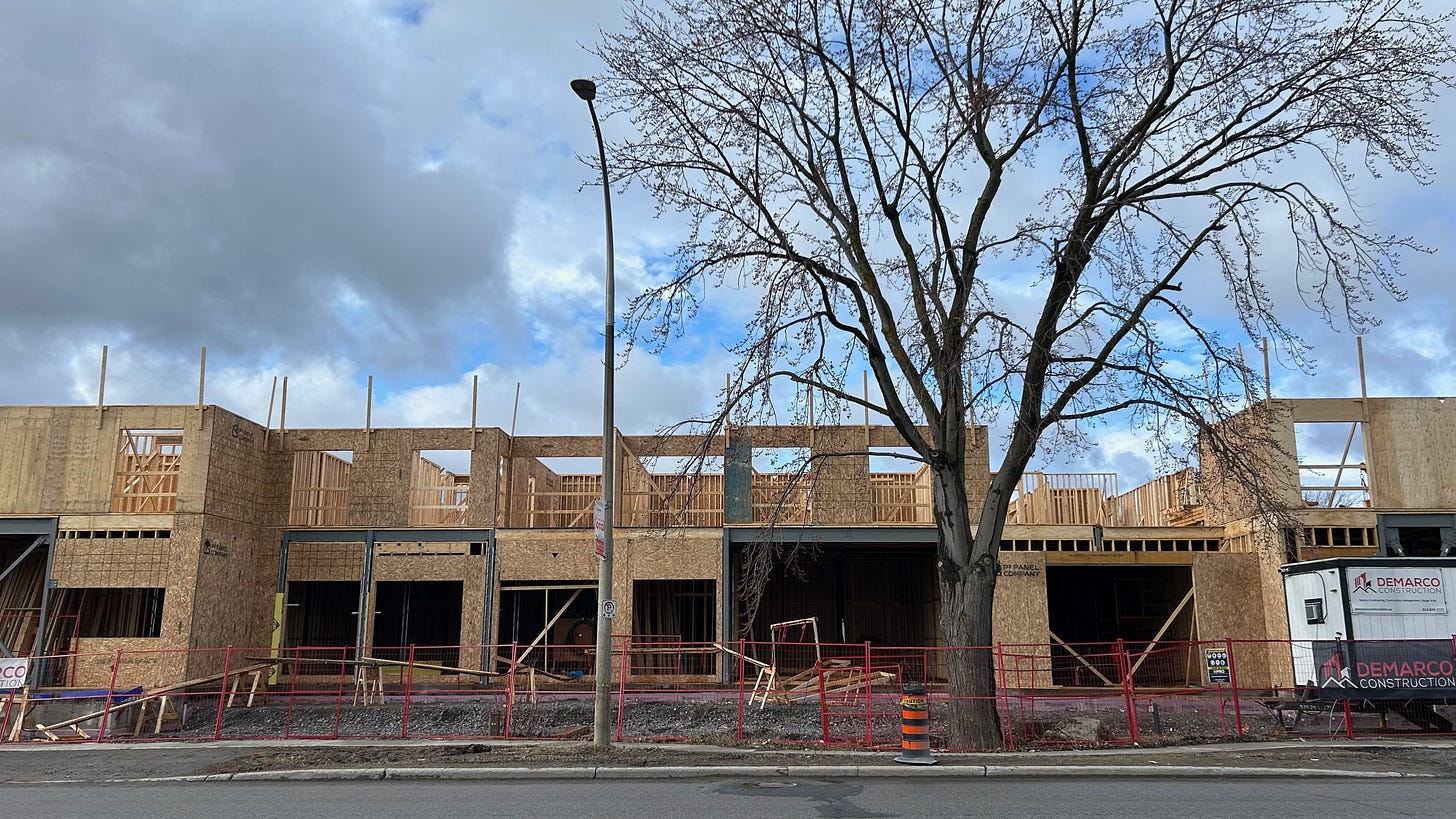 The wood frame of the first and second floor of an apartment building. A lamp post and tree are in the foreground. Construction fencing all around the site.