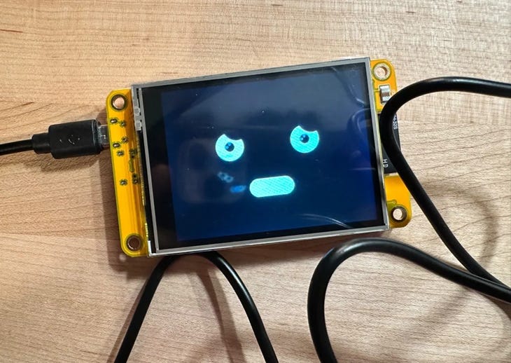 Photo of a small yellow ESP32 display showing a blue cartoon face with wide eyes and a frown on a dark screen, lying on a wooden desk with connected cables.