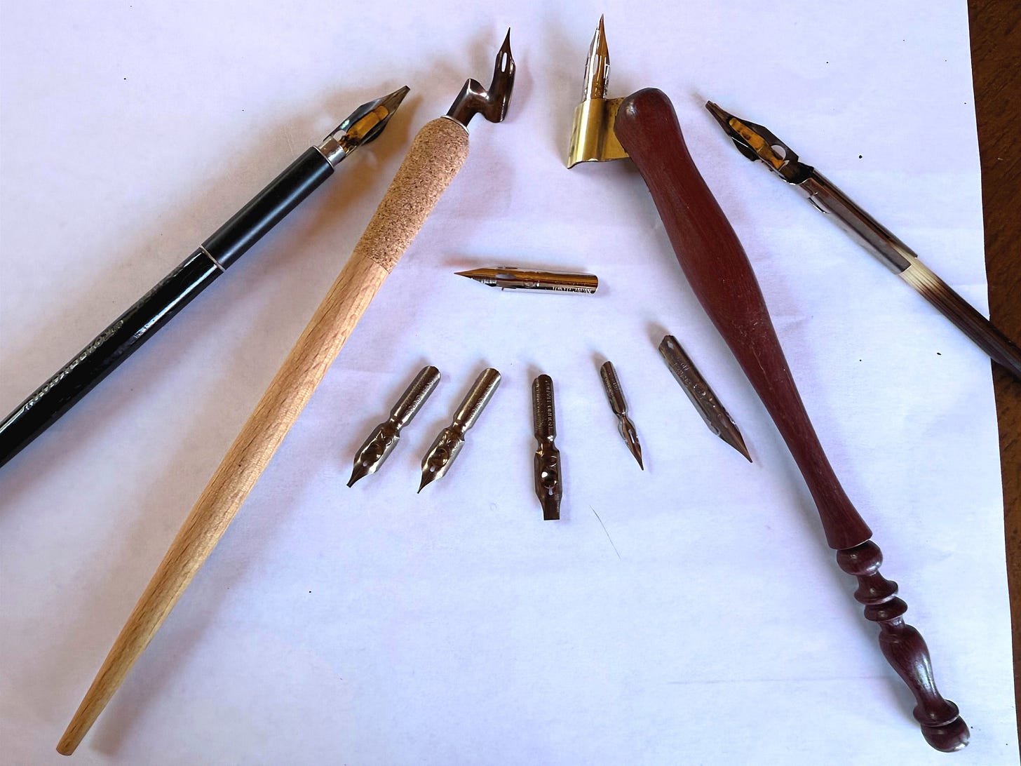 A selection of calligraphy dip pens and metal nibs arranged on a sheet of white paper.