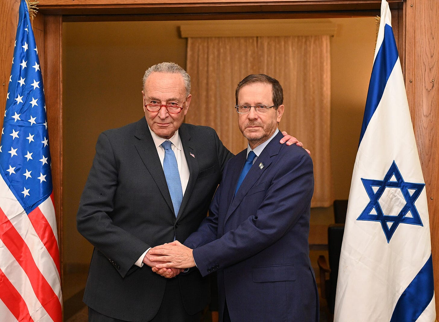 two white men in dark suits shaking hands; on their right an american flag, on left Israeli flag