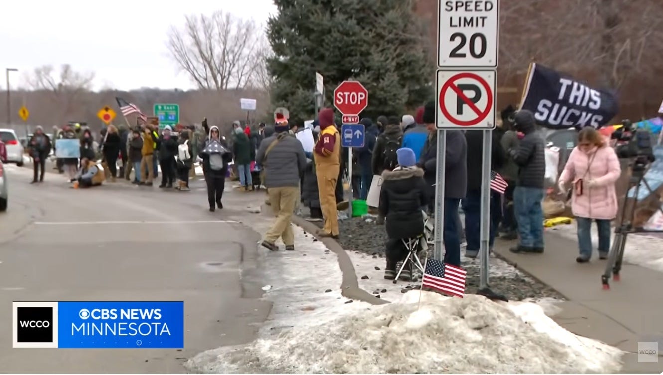 Video screenshot of 20-30 warmly dressed protesters on sidewalks outside a federal building, behind a pile of dirty snowplowed days ago. They carry signs and US flags. To one side a protester has unfurled a large black flag with white letters reading 'THIS SUCKS'