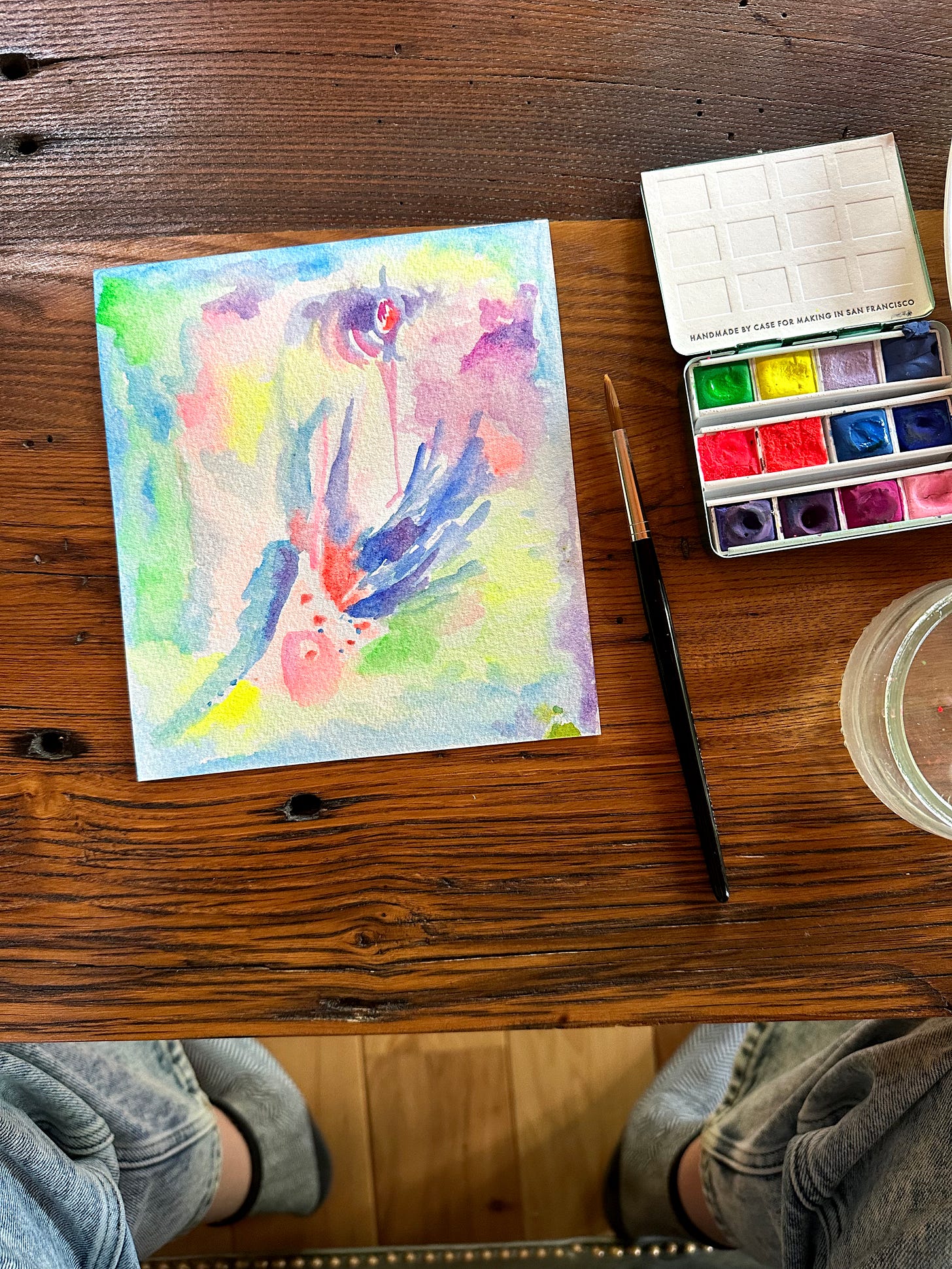 An image looking down at a wooden surface with a painting full of abstract vivid neon colors and shapes, pinks, blues, yellows, greens...and to the right a small palette of bright paints and a paintbrush, and a little corner of a water jar. 