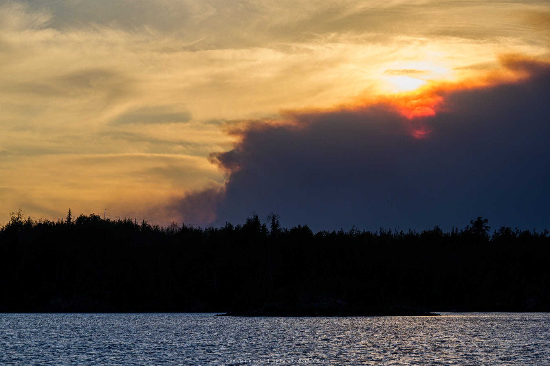 The setting sun glows deep red through a massive plume of wildfire smoke rising above a forested shoreline and lake in northeastern Minnesota.