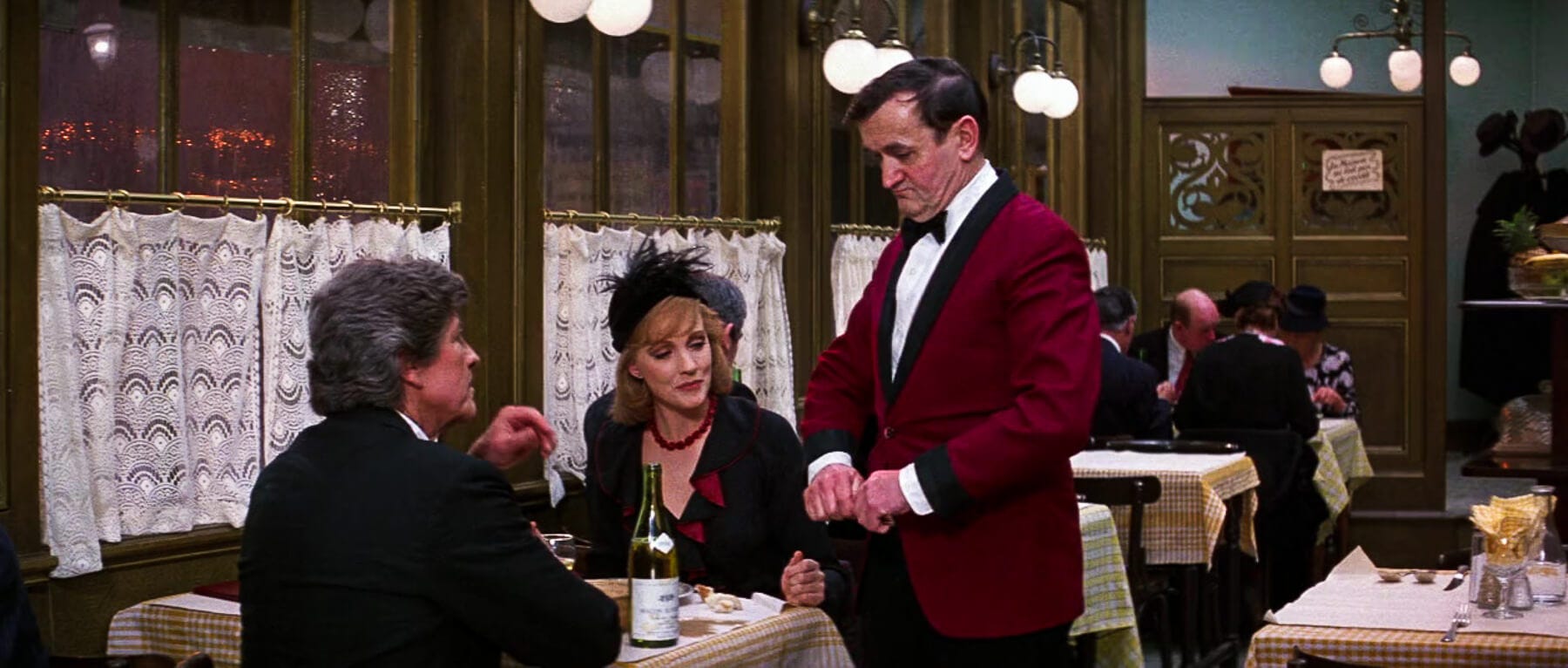 Graham Stark, stone-faced in a red waiter jacket, delivers attitude to a disgruntled customer at a bustling bistro table—wine bottle mid-pour. Graham Stark, stone-faced in a red waiter jacket, delivers attitude to a disgruntled customer at a bustling bistro table—wine bottle mid-pour.