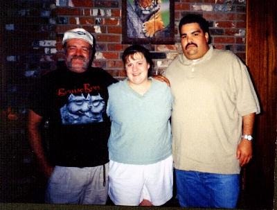 Photograph from 1996 showing the author standing between her birth father, Leamon (left), and half-brother, Justin (right), in front of a brick wall. All three are smiling slightly, arms around each other in a casual family pose.
