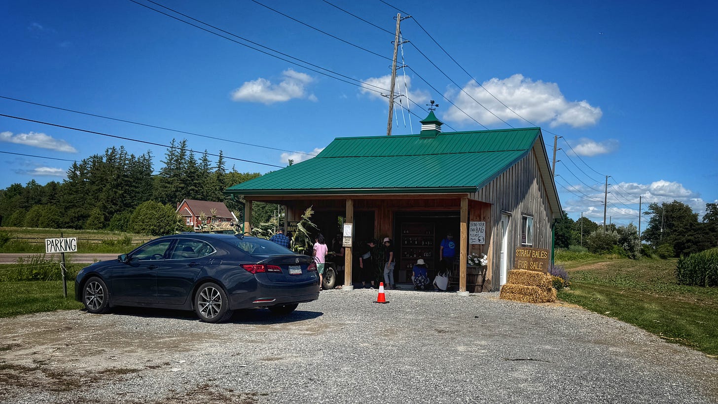 The parking lot and corn stand at Sweet Ridge Farms. There's a black car parked in front of a small corn stand building with a peaked green roof. Some people are in front of the building. There's a sign that reads, "Straw Bales for Sale." The parking lot and corn stand at Sweet Ridge Farms. There's a black car parked in front of a small corn stand building with a peaked green roof. Some people are in front of the building. There's a sign that reads, "Straw Bales for Sale."