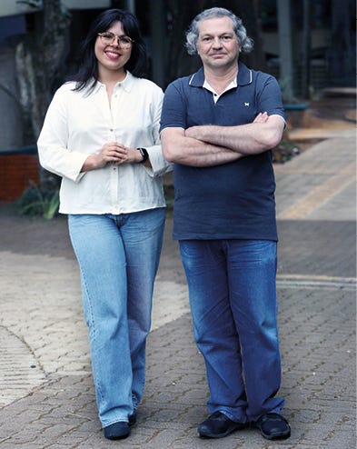 Duas pessoas em pé lado a lado em um espaço externo com piso de tijolos, uma vestindo blusa branca de manga comprida e calça jeans azul, a outra vestindo polo azul-marinho com gola branca e calça jeans azul, ambas sorrindo para a câmera, com edifícios desfocados ao fundo. Duas pessoas em pé lado a lado em um espaço externo com piso de tijolos, uma vestindo blusa branca de manga comprida e calça jeans azul, a outra vestindo polo azul-marinho com gola branca e calça jeans azul, ambas sorrindo para a câmera, com edifícios desfocados ao fundo.