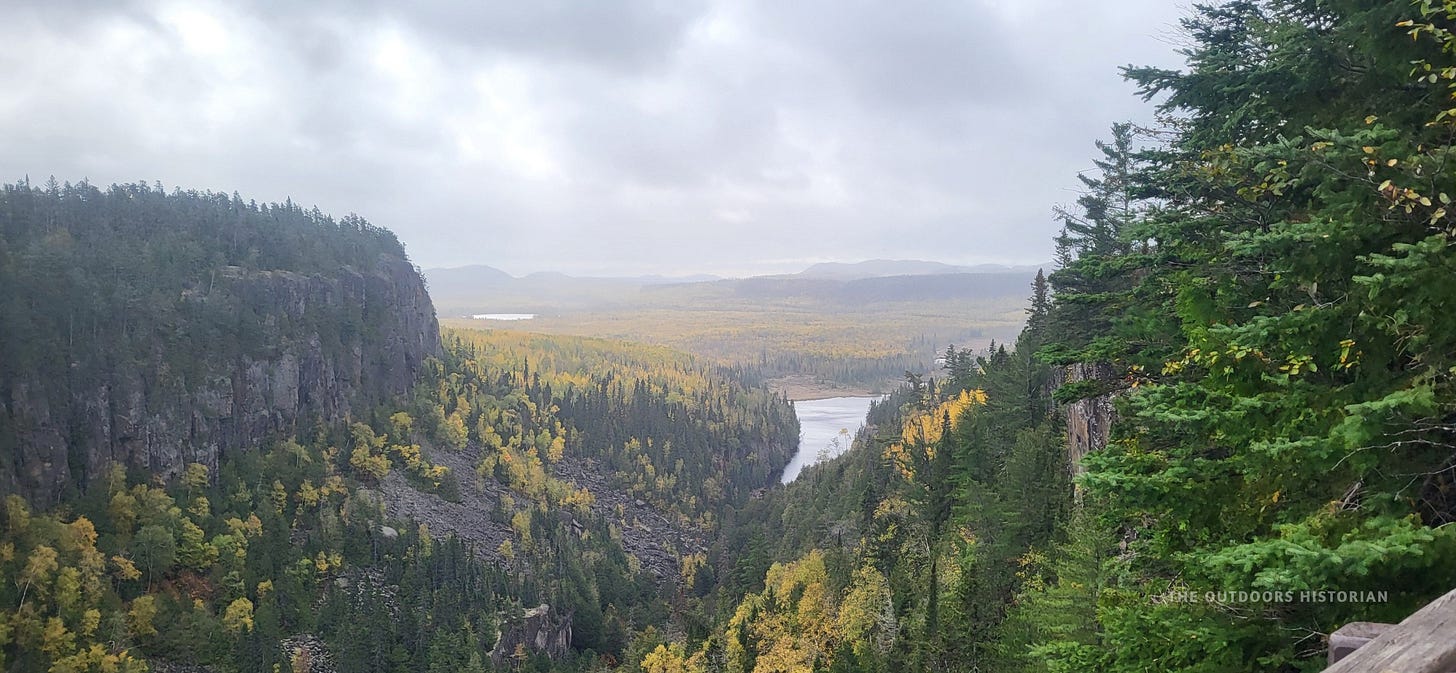 A high-angle view looking down into a massive rock canyon. The canyon floor is in deep shadow, while the rim is lined with trees showing muted fall colors of yellow and orange against a cloudy grey sky.