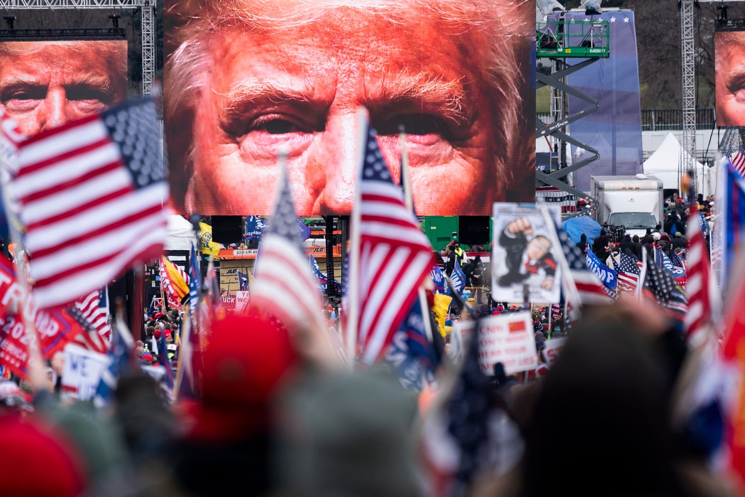 A shot of the Ellipse stage from the crowd; above US flags and homemade signs featuring Trump as a superhero, loom three giant video screens, each with a high contrast image of Trump’s eyes gazing out at the crowd. A shot of the Ellipse stage from the crowd; above US flags and homemade signs featuring Trump as a superhero, loom three giant video screens, each with a high contrast image of Trump’s eyes gazing out at the crowd.