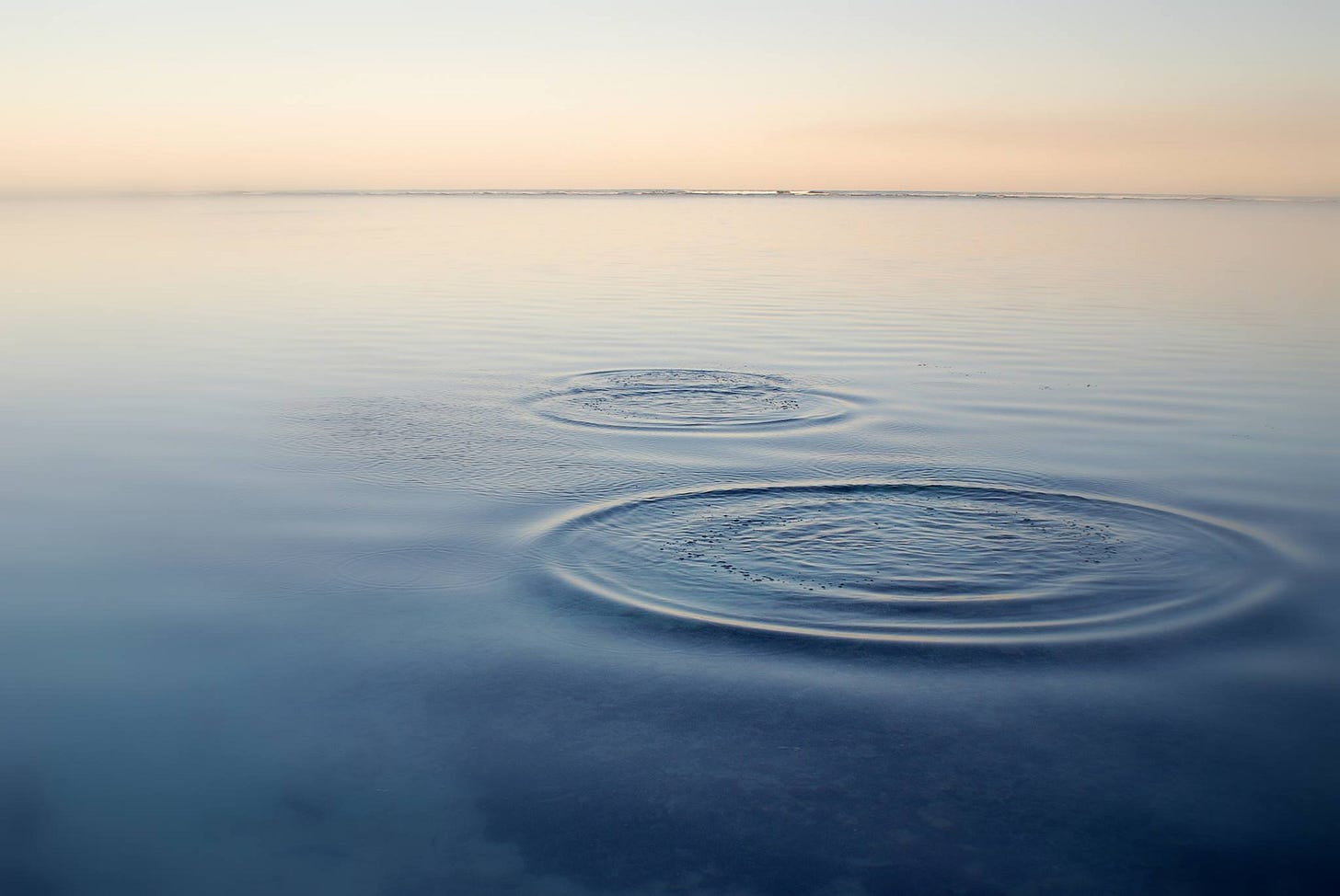 A calm sea at dusk with two concentric ripples widening slowly across the water’s surface.