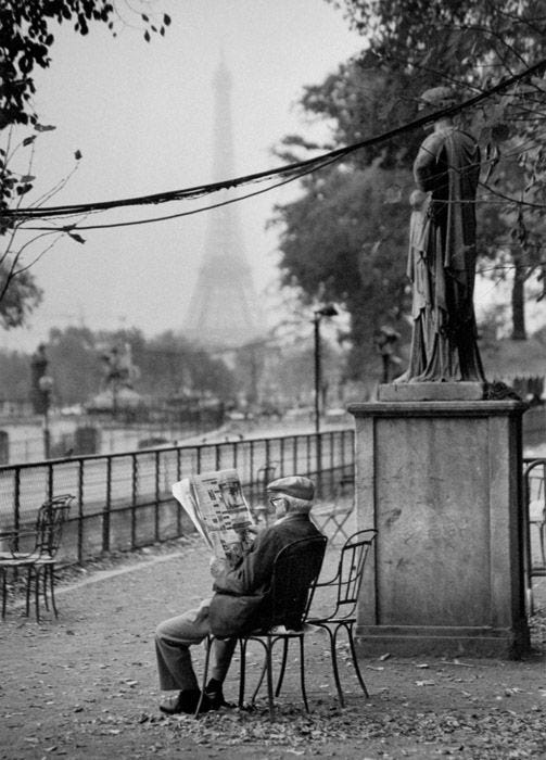 This may contain: a man sitting on a chair reading a newspaper in front of the eiffel tower