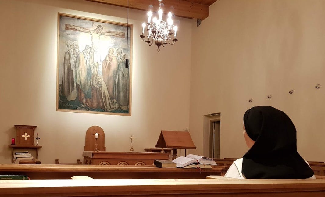 Ett kapell med ett enkelt tabernakel i trä längst framme till vänster. Till höger ser man en dominikansyster sitta. På väggen ovanför altaret finns en altartavla med Kristus på korset och sörjande människor omkring korsets fot. Men kring Kristusfiguren syns en ljusrundel som nästan täcker honom, så att man där kan se både oblaten och den korsfäste.