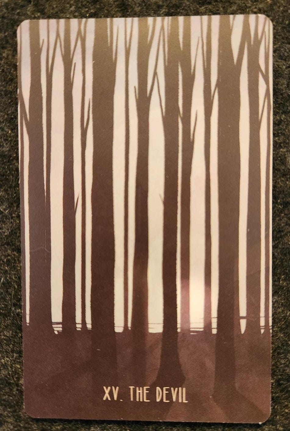 A tarot card labelled XV. The Devil. A row of tall, dark, leafless trees in front of a grey sky with a subtle, muted, sun peeking through.