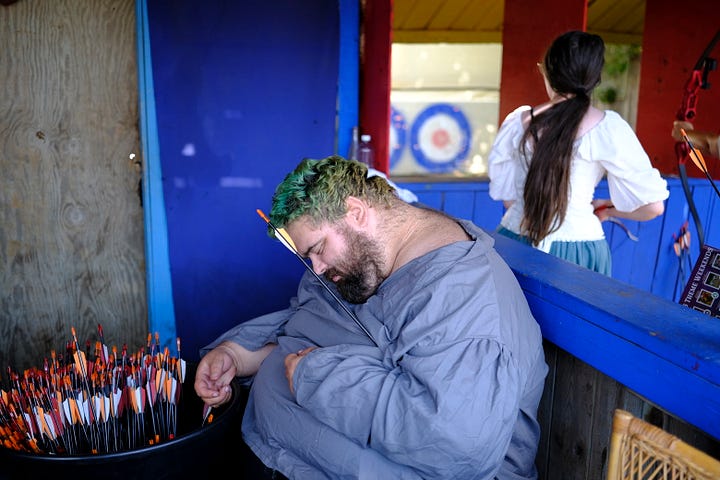 Bards singing in a tavern wearing medieval garb, an elf and wizard surveying goods at a shop, fire-twirler show, Geralt and another warrior deep in conversation, a man playing dead with an arrow under his armpit, guests walking toward the camera, a woman with a horse head looking over a table.