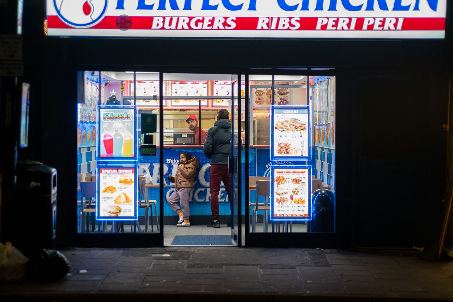 Fast food shop at night, lit by fluorescent menus. Inside a girl in a brown jacket looks out, while a man faces the server in red polo and cap.