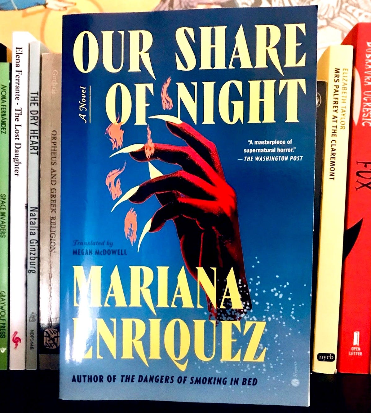 A copy of Mariana Enriquez's Our Share of Night standing upright, against a row of other books in the background.