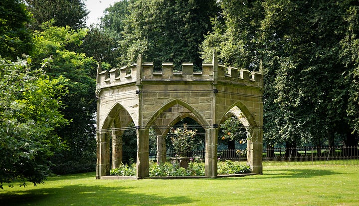 A stone gazebo with Gothic arches and battlements stands in a lush, green garden surrounded by tall trees and vibrant foliage.