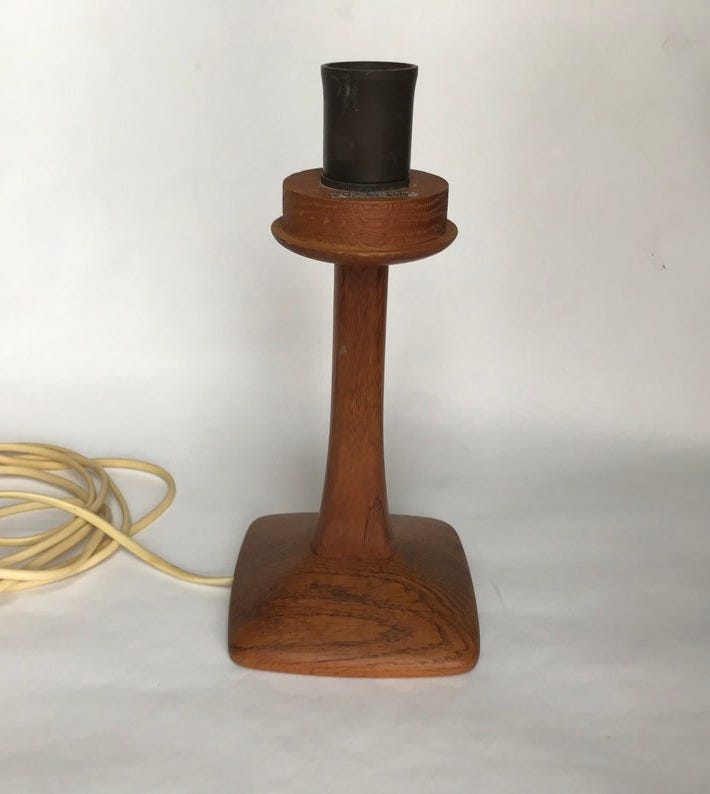 May include: A wooden table lamp with a square base and a cylindrical top. The lamp has a brown finish and a dark metal socket. A cream-colored electrical cord with a white plug is visible to the left of the lamp.