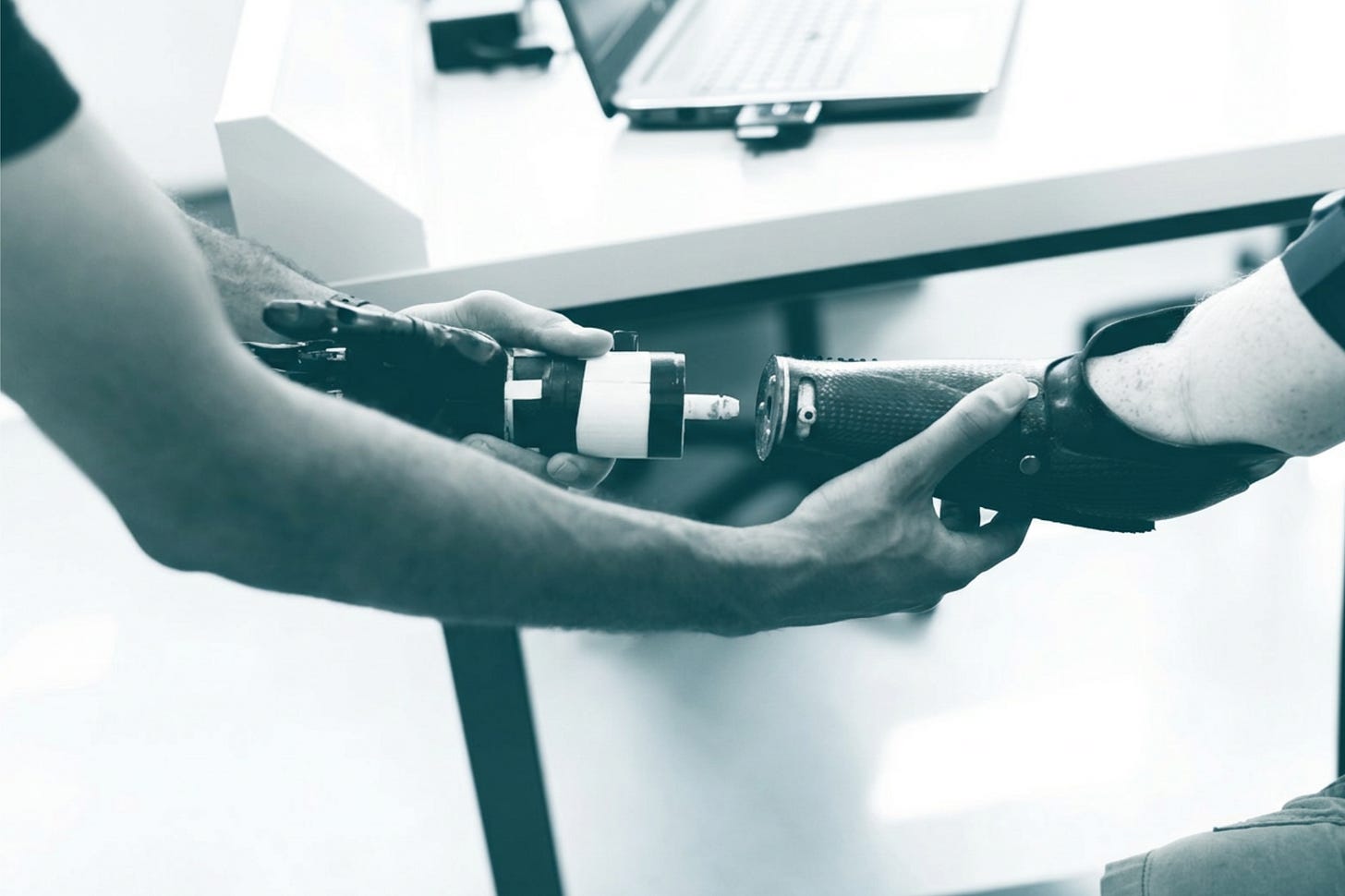 Two people's hands fitting a prosthetic arm component to a carbon-fiber socket on a person's forearm, with a laptop on a white desk behind them. Two people's hands fitting a prosthetic arm component to a carbon-fiber socket on a person's forearm, with a laptop on a white desk behind them.
