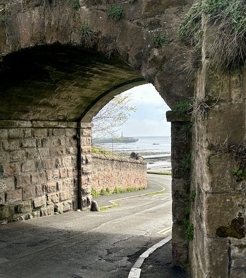 An stone archway through which a narrow road runs through with the sea and a lighthouse in the distance at the end of a causeway. An stone archway through which a narrow road runs through with the sea and a lighthouse in the distance at the end of a causeway.
