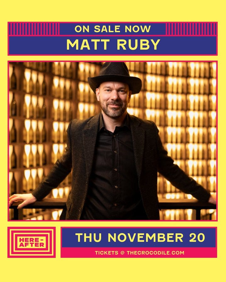 Vibrant promotional poster with yellow background and purple red borders. Central image shows Matt Ruby a man with short hair wearing a black fedora hat dark gray suit jacket black shirt and pants standing with arms outstretched in front of a wall of numerous clear glass wine bottles illuminated with warm light. Text overlays include On sale now in white on purple banner Matt Ruby in large white letters on blue banner Tickets at thecrocodile.com in pink on yellow banner Here After logo in pink and blue and Thu November 20 Tickets at thecrocodile.com in blue and pink.