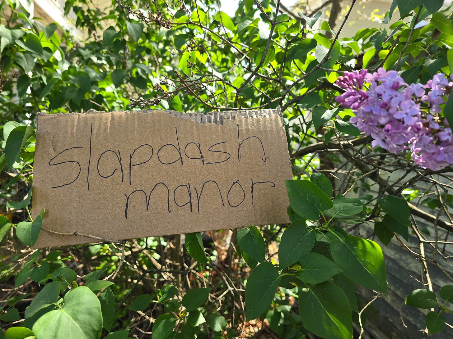 A handwritten sign on a bit of cardboard is nestled into a lilac bush. It reads "Slapdash Manor."