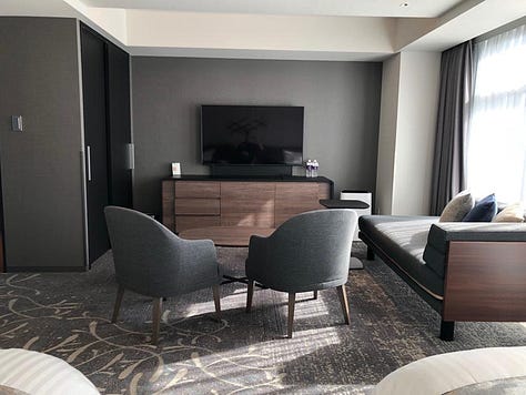 A modern and spacious hotel room with twin beds, a seating area with armchairs and a chaise lounge, a flat-screen TV, and large windows with sheer curtains. The accessible bathroom features grab bars, a handheld showerhead, and a shower chair.