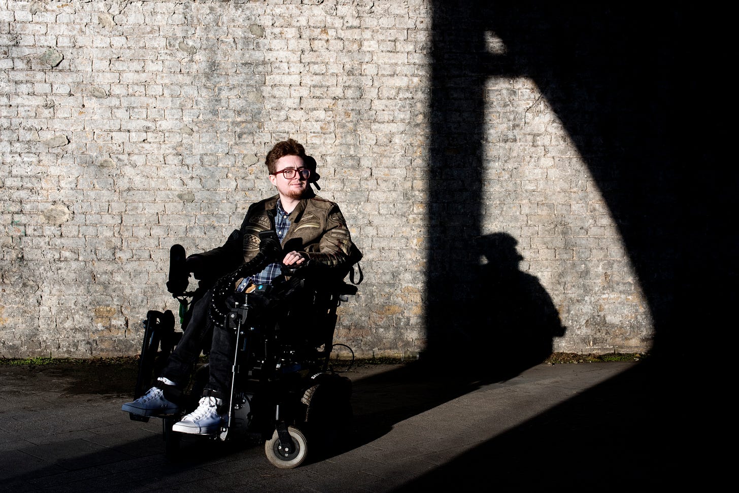 Jamie, a white electric wheelchair user sis against a sunsoaked wall, crisp shadows in the background
